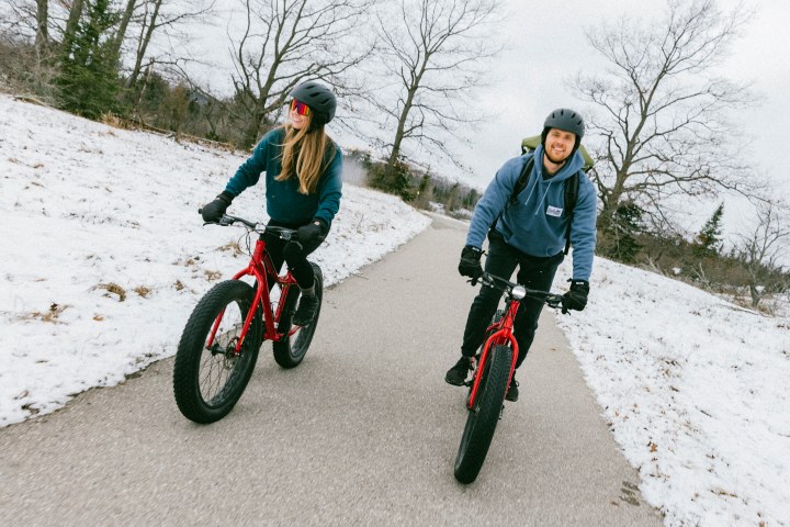 Two people biking on a snowy path, wearing helmets and winter gear.