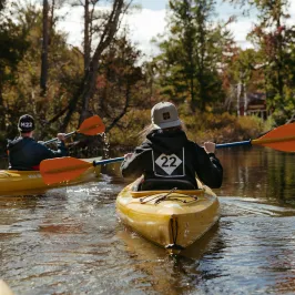 Two kayakers paddle in yellow kayaks through a calm river surrounded by trees.