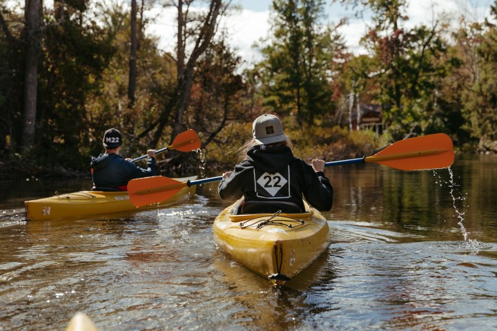 Two kayakers paddle in yellow kayaks through a calm river surrounded by trees.