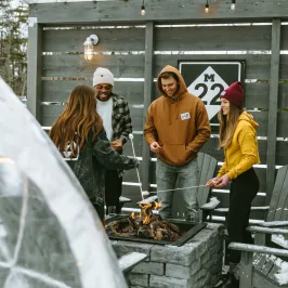 Four people roasting marshmallows over a fire pit near a wooden fence with lights.