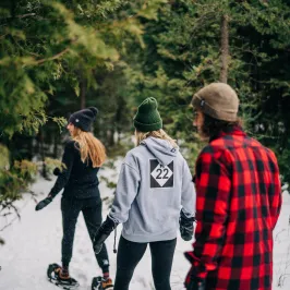 Three people snowshoeing through a snowy forest, wearing winter clothing and beanies.