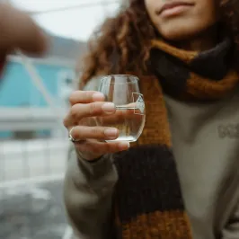 Person holding a glass of clear liquid, wearing a brown and yellow scarf outside.