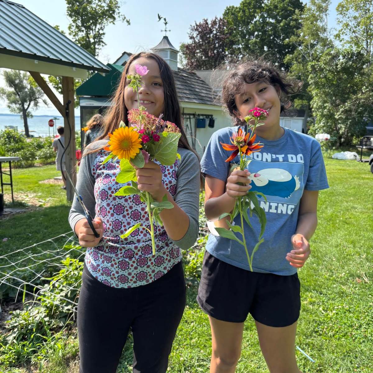 Two people holding flowers, standing outdoors on a grassy area near buildings.