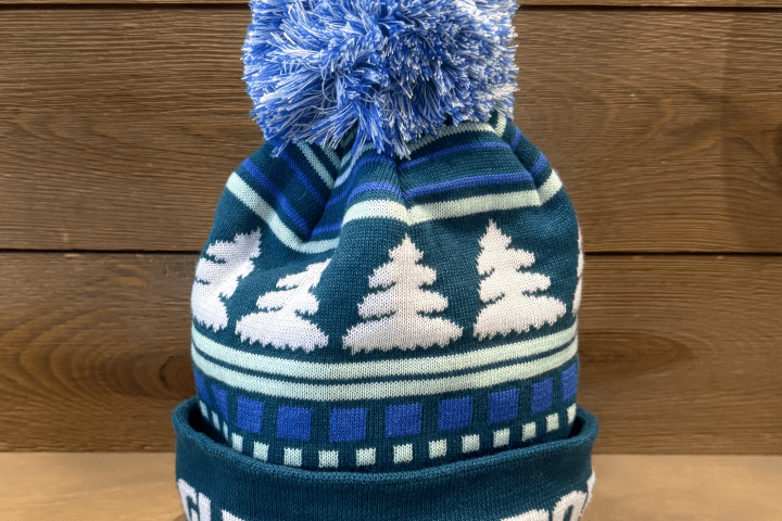 Blue knit beanie with white tree pattern and pom-pom on top on a wooden background.