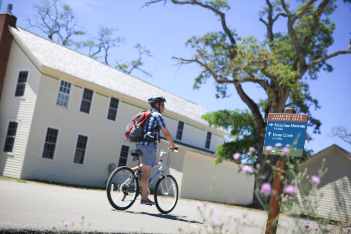 Person with bike near beige building and tree, sign indicates Marinelife Museum and Dune Climb directions.