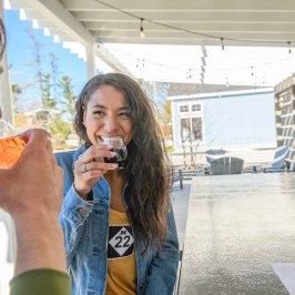 Two people smiling and drinking beverages on a patio with string lights overhead.